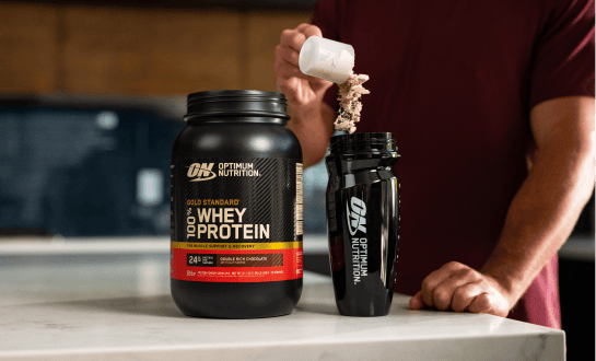 Person pouring whey protein powder from a scoop into a shaker bottle beside a container of Optimum Nutrition Gold Standard 100% Whey Protein on a kitchen countertop.